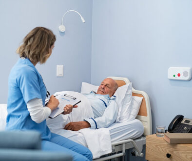 A Nurse Talking With a Senior Patient While Writing on a Clipboard Sitting on the Edge of a Hospital Bed How Long Can You Stay in Inpatient Hospice