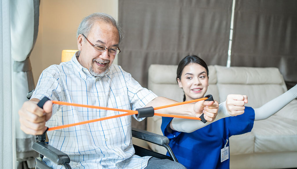 A Senior Man Sitting in a Wheelchair Beside a Female Occupational Therapist Using a Resistance Band During In-Home Occupational Therapy