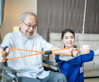 A Senior Man Sitting in a Wheelchair Beside a Female Occupational Therapist Using a Resistance Band During In-Home Occupational Therapy