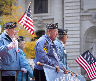 Three Veterans Standing on a Float Holding American Flags During a Parade Veterans in Hospice