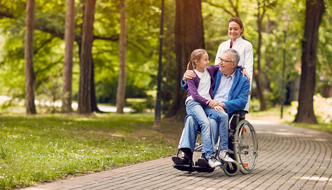 A Senior Man in a Wheelchair With His Granddaughter Sitting on His Lap and a Caregiver Standing Behind Them Hospice Donations