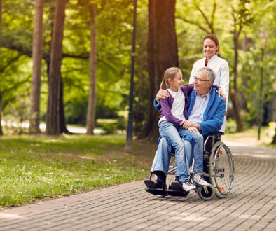 A Senior Man in a Wheelchair With His Granddaughter Sitting on His Lap and a Caregiver Standing Behind Them Hospice Donations