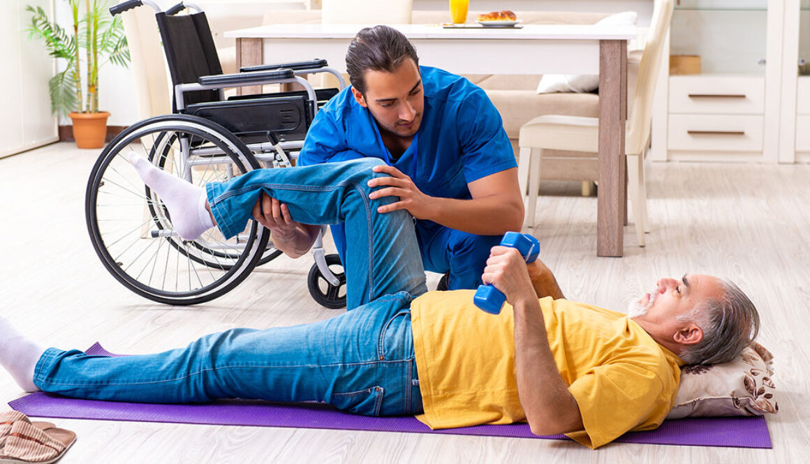 A Young Male Physical Therapist Helping A Senior Man Laying On His Back Stretch His Hip With At-Home Physical Therapy