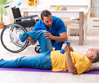 A Young Male Physical Therapist Helping A Senior Man Laying On His Back Stretch His Hip With At-Home Physical Therapy