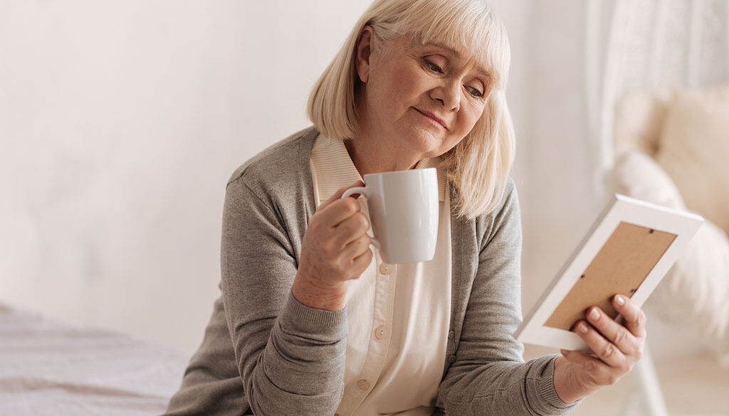 An Older Woman Holding A Coffee Mug Looking At A Picture Grieving The Loss Of A Spouse