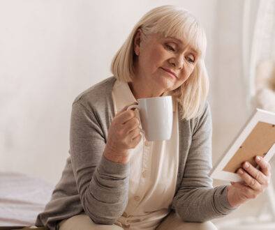 An Older Woman Holding A Coffee Mug Looking At A Picture Grieving The Loss Of A Spouse