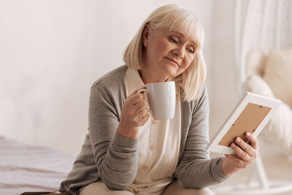 An Older Woman Holding A Coffee Mug Looking At A Picture Grieving The Loss Of A Spouse